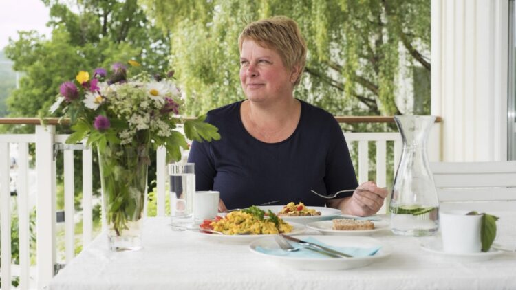 Woman eating a colorful brunch outside on a white table decorated with flowers, on a white porch in green surroundings.